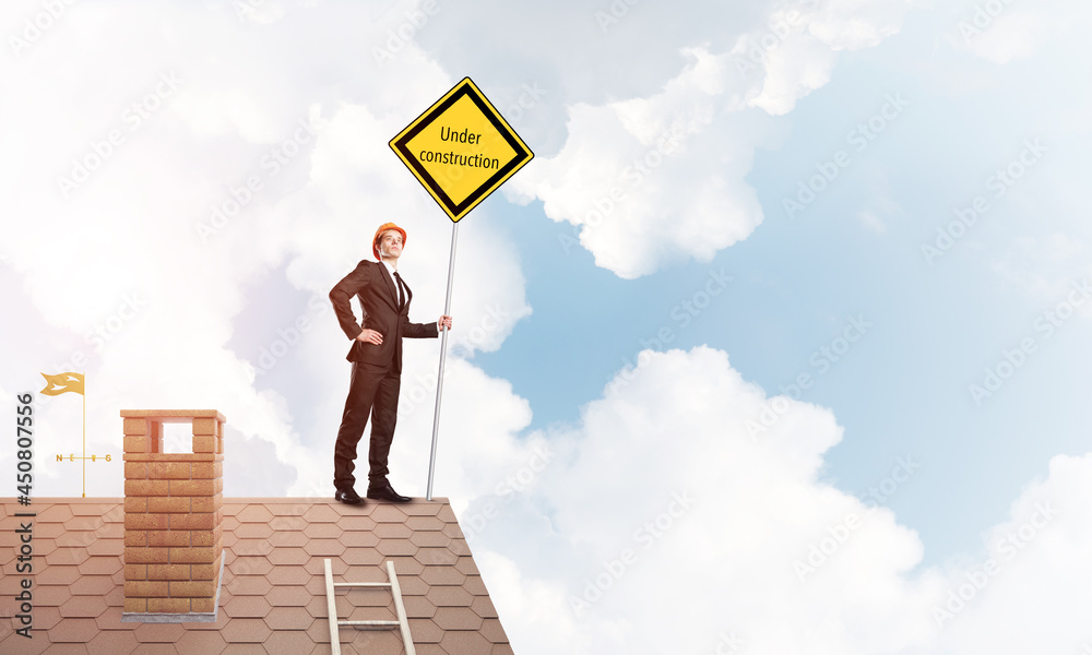 Young businessman on house brick roof holding yellow signboard. Mixed media Young businessman on house brick roof holding yellow signboard. Mixed media
