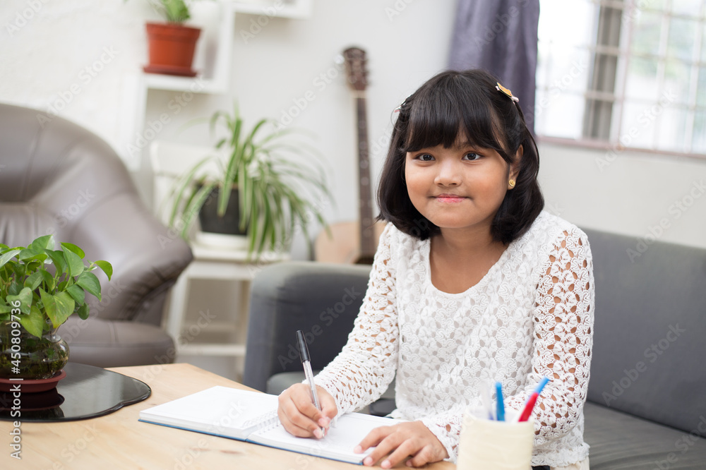 Happy little girl sitting and writing her homework with pen in a notebook at home. Smiling child dressed in white, holding pen ready to write. 