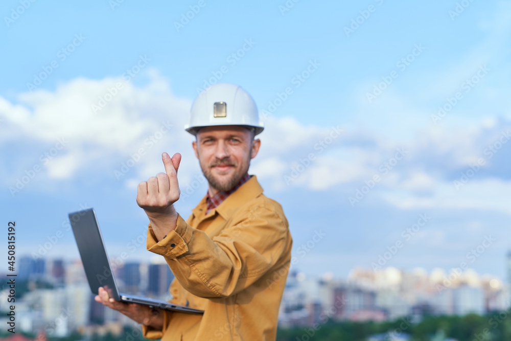 Bearded supervisor, foreman or construction inspector looking at camera and making Korean heart gesture. Contractor in hardhat and orange jacket holding computer laptop in front of sunny day blue sky Bearded supervisor, foreman or construction inspector looking at camera and making Korean heart gesture. Contractor in hardhat and orange jacket holding computer laptop in front of sunny day blue sky