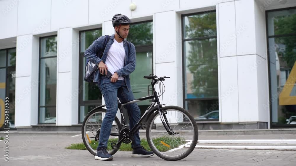 Young caucasian courier male delivering food with transportation thermo backpack looking for delivery address using phone standing in city street. Deliveryman pushing bicycle and riding to client. Young caucasian courier male delivering food with transportation thermo backpack looking for delivery address using phone standing in city street. Deliveryman pushing bicycle and riding to client.