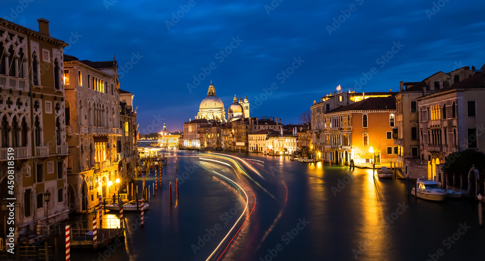 Beautiful view in the night at Cityscape image of Grand Canal in Venice, with Santa Maria della Salute Basilica reflected in calm sea. Lights of passenger boat on the water.