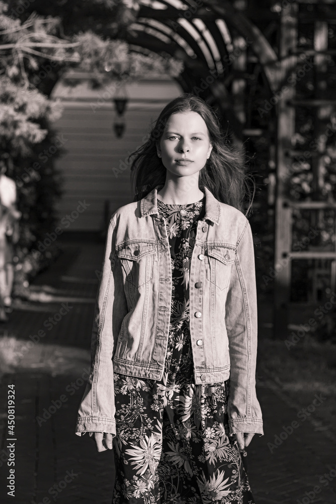 Monochrome portrait of a young girl in the park. The sun's rays illuminate the face of a girl standing in the shade. She is wearing a silk floral dress and a denim jacket.Natural beauty without makeup Monochrome portrait of a young girl in the park. The sun's rays illuminate the face of a girl standing in the shade. She is wearing a silk floral dress and a denim jacket.Natural beauty without makeup