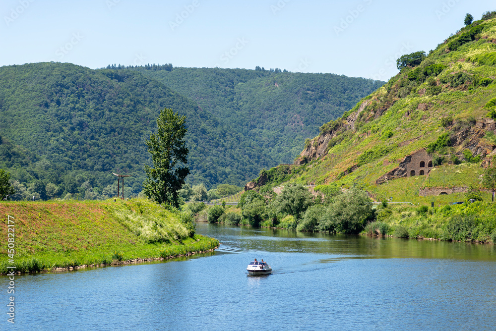 Two men are sailing a motorboat on a calm river between the hills covered with forest.