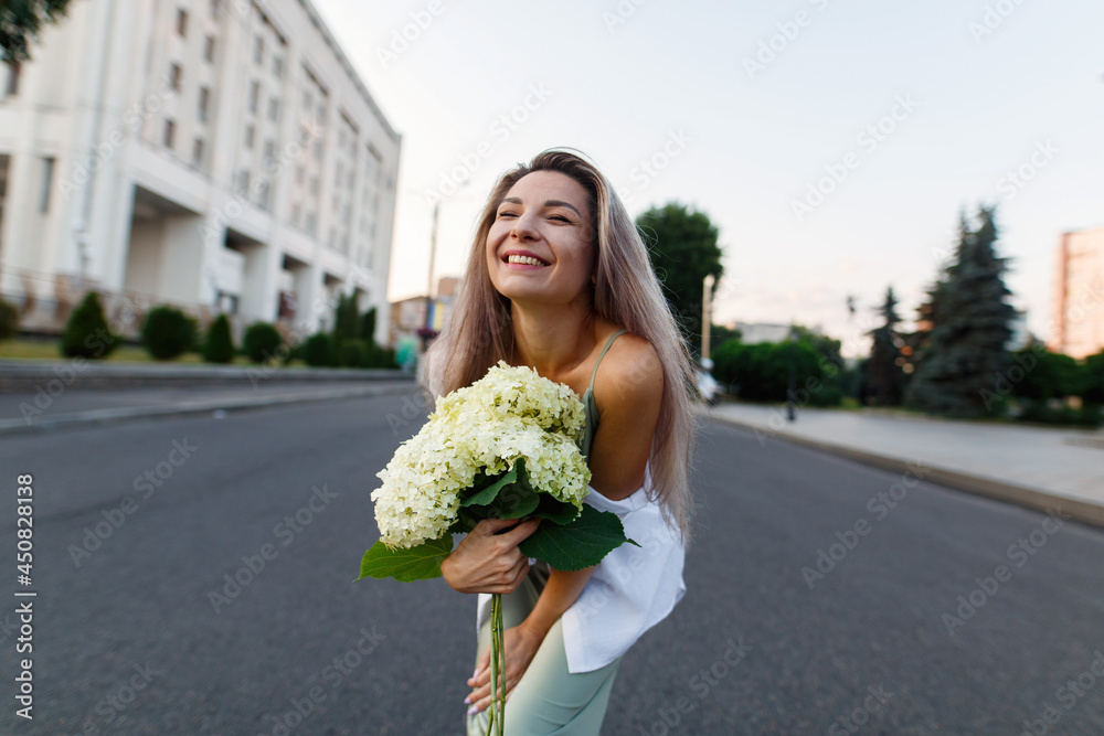 Fototapeta premium portrait a happy emotional woman in street style. smiling girl in dress with flowers walking on city street in summer morning. sexy blonde woman with bouquet hydrangea outdoors on road town.