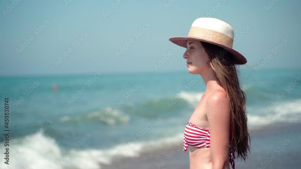 Portrait in profile of charming brunette woman in summer hat pensively looking in distance on amazing rough waves background, attractive female in colorful bikini enjoying sunlight, vacations, relax Portrait in profile of charming brunette woman in summer hat pensively looking in distance on amazing rough waves background, attractive female in colorful bikini enjoying sunlight, vacations, relax
