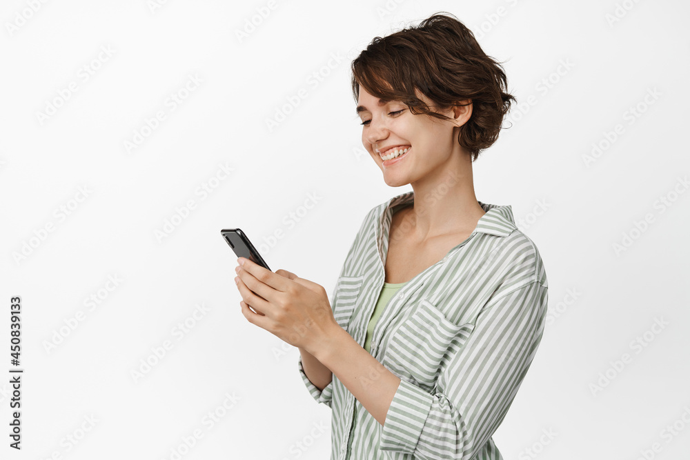 Portrait of smiling girl chatting on mobile phone, using smartphone cellular, standing in casual outfit against white background Portrait of smiling girl chatting on mobile phone, using smartphone cellular, standing in casual outfit against white background