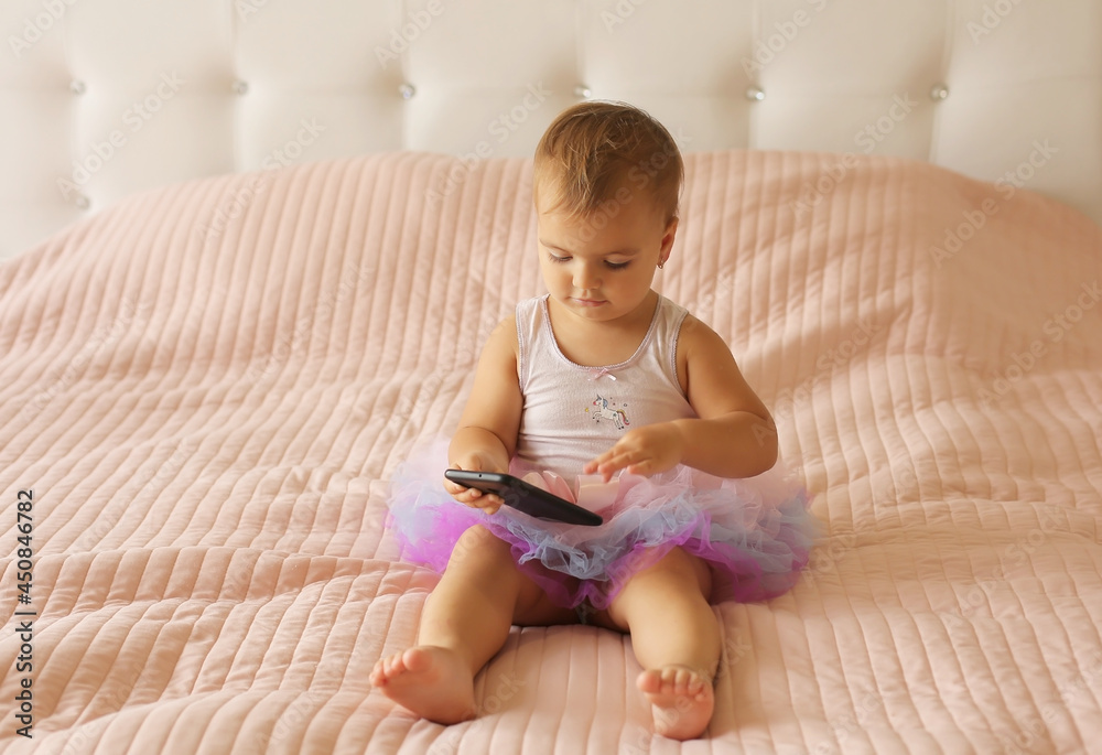 a little happy beautiful girl is sitting on a pink blanket in a skirt and holding a phone in her hands a little happy beautiful girl is sitting on a pink blanket in a skirt and holding a phone in her hands