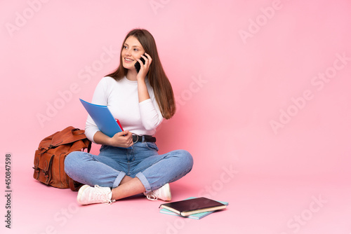 Teenager caucasian student girl sitting on the floor isolated on pink background keeping a conversation with the mobile phone