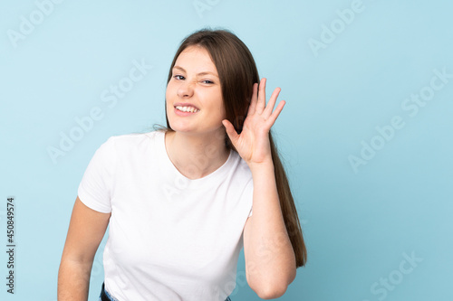 Teenager caucasian girl isolated on blue background listening to something by putting hand on the ear © luismolinero