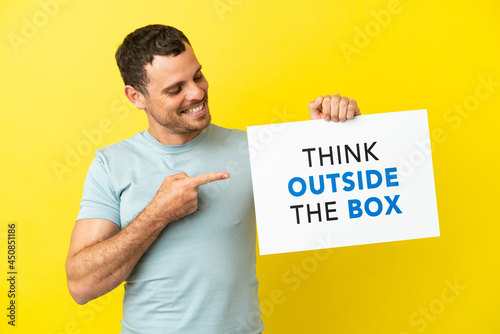 Brazilian man over isolated purple background holding a placard with text Think Outside The Box and  pointing it © luismolinero