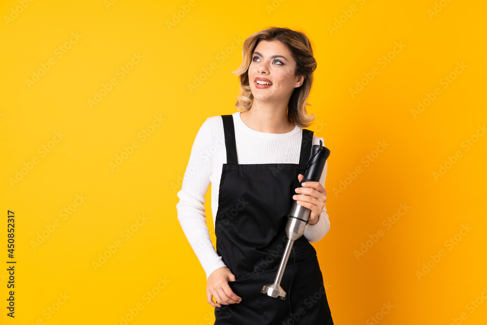 Girl using hand blender isolated on yellow background thinking an idea while looking up Girl using hand blender isolated on yellow background thinking an idea while looking up