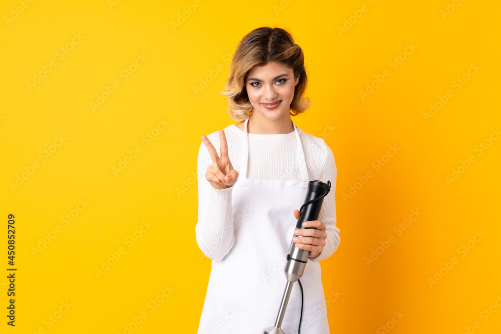 Girl using hand blender isolated on yellow background smiling and showing victory sign