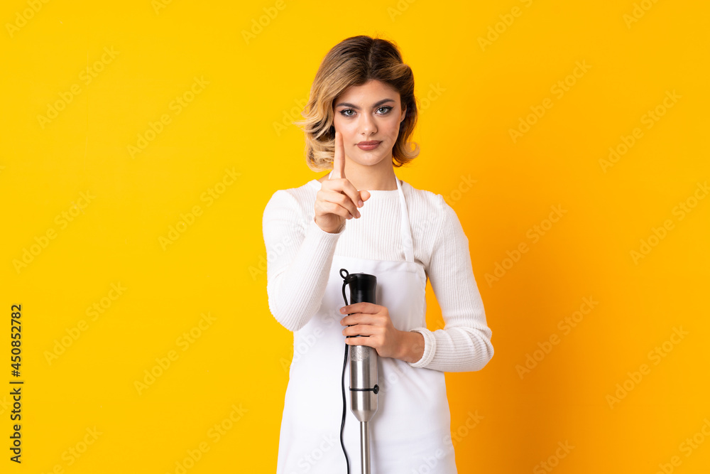 Girl using hand blender isolated on yellow background showing and lifting a finger Girl using hand blender isolated on yellow background showing and lifting a finger