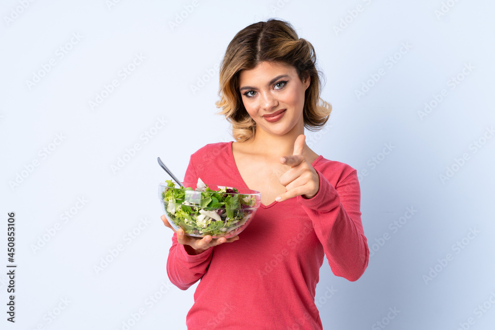 Teenager woman with salad isolated on blue background points finger at you with a confident expression Teenager woman with salad isolated on blue background points finger at you with a confident expression