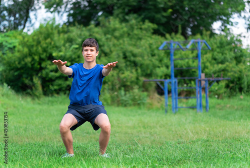 teenage boy exercising outdoors, sports ground in the yard, he squats and does a warm-up, healthy lifestyle