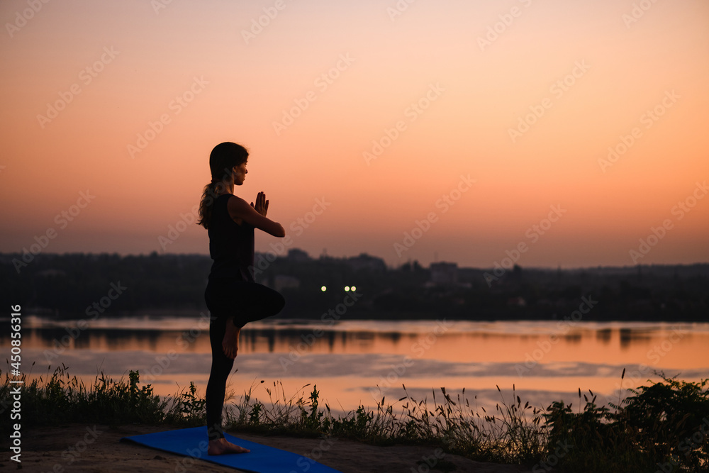 Fototapeta premium Morning sunrise at water and city horizon with healthy woman standing in balance yoga Vrksasana tree position