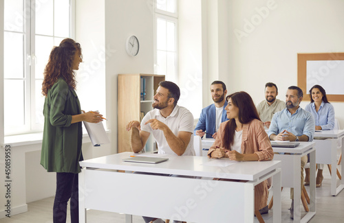 College teacher having class with group of interested adult male and female business course students. People sitting at tables in classroom, discussing topic of success, asking or answering questions