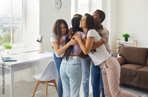 'Long time no see' or 'Congratulations, girl, you did it' scene: Diverse group of happy excited young women hugging their friend as they meet at a friendly get together, gathering or fun party at home