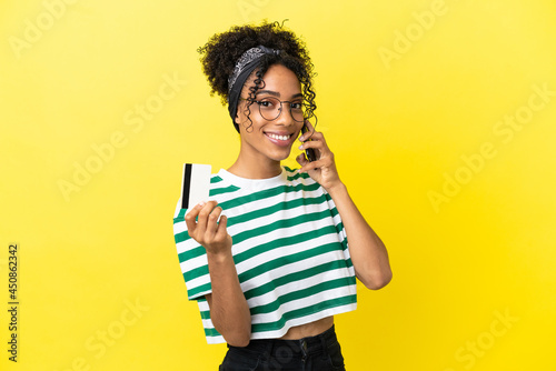 Young african american woman isolated on yellow background keeping a conversation with the mobile phone and holding a credit card