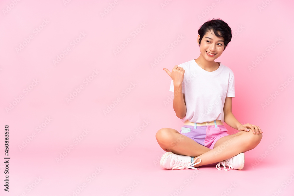 Young Vietnamese woman with short hair sitting on the floor over isolated pink background pointing to the side to present a product Young Vietnamese woman with short hair sitting on the floor over isolated pink background pointing to the side to present a product