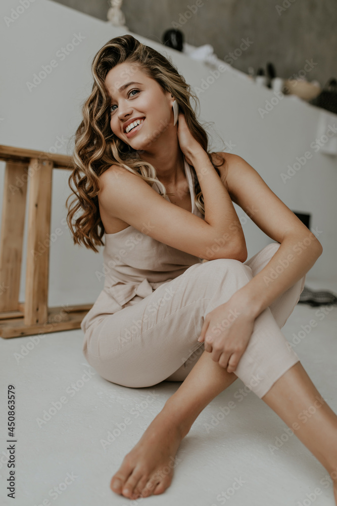 Pretty blonde curly woman in trendy beige jumpsuit smiles sincerely and sits on white floor in cozy light bathroom. Pretty blonde curly woman in trendy beige jumpsuit smiles sincerely and sits on white floor in cozy light bathroom.