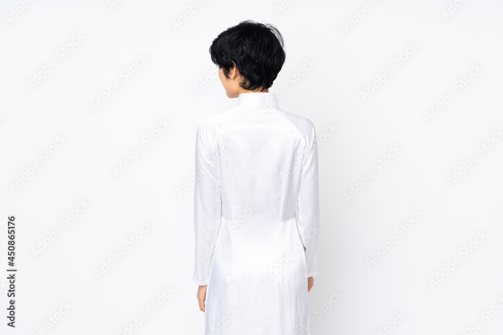 Young Vietnamese woman with short hair wearing a traditional dress over isolated white background in back position and looking side Young Vietnamese woman with short hair wearing a traditional dress over isolated white background in back position and looking side