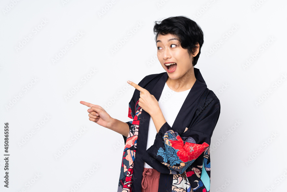 Young Vietnamese woman with short hair wearing a traditional dress over isolated white background surprised and pointing side Young Vietnamese woman with short hair wearing a traditional dress over isolated white background surprised and pointing side