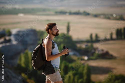 Fototapeta Naklejka Na Ścianę i Meble -  Guy looking at binoculars in hill. man in t-shirt with backpack. Young Caucasian man during hike in valley landscape