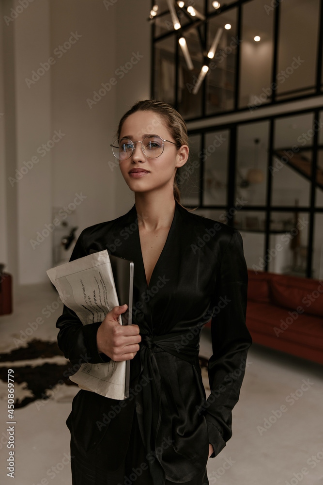 Pretty elegant woman in eyeglasses, black silk stylish home suit poses in living room, looks into camera and holds laptop and newspaper. Pretty elegant woman in eyeglasses, black silk stylish home suit poses in living room, looks into camera and holds laptop and newspaper.