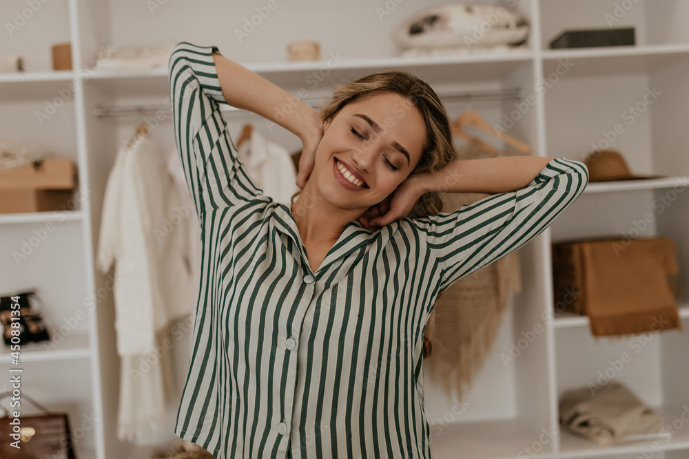 Happy young woman in striped shirt stretches at home. Blonde curly lady in silk blouse smiles sincerely in dressing room. Happy young woman in striped shirt stretches at home. Blonde curly lady in silk blouse smiles sincerely in dressing room.