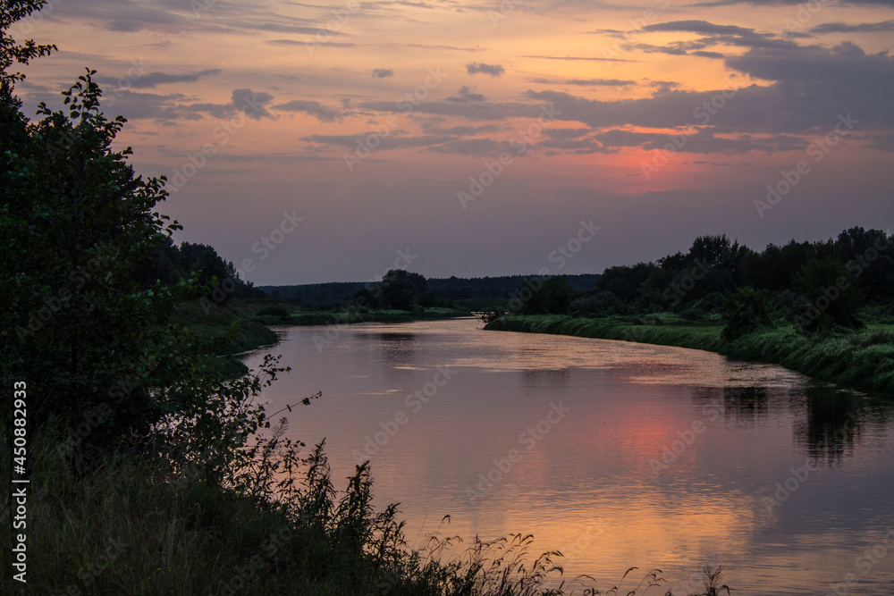 A beautiful landscape - a dramatic sunset sky and a river with reflection and banks with silhouettes of grass and trees and a space to copy