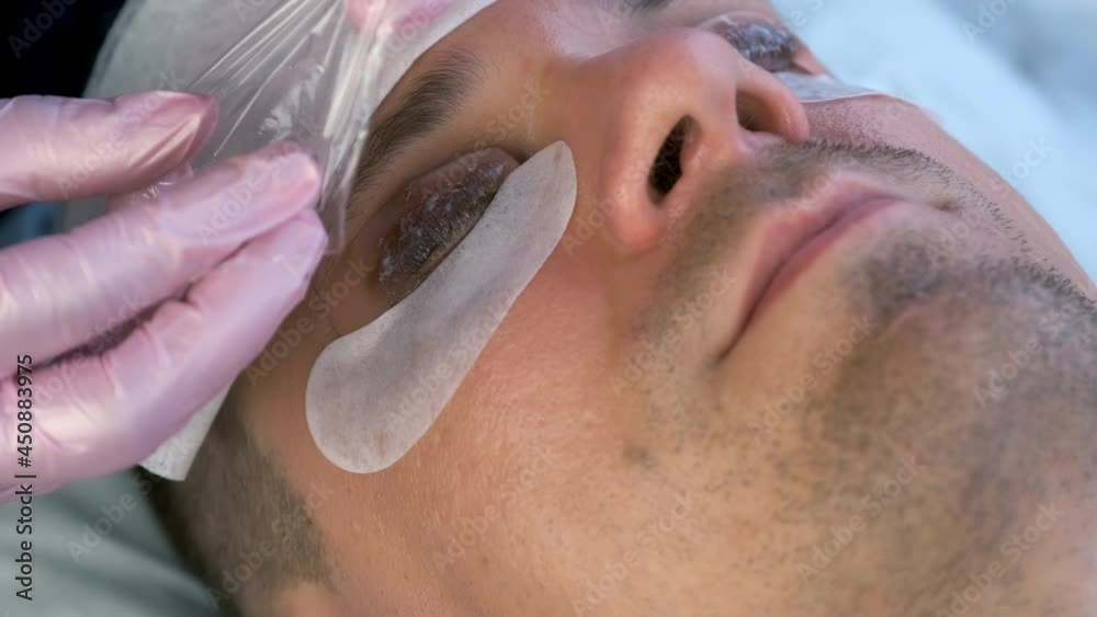 Beautician covered man's lashes with file, pads and towel, lift eyelashes procedure. Man with solution for lamination on lashes, face closeup. Cosmetologist making lash lifting in cosmetology clinic. Beautician covered man's lashes with file, pads and towel, lift eyelashes procedure. Man with solution for lamination on lashes, face closeup. Cosmetologist making lash lifting in cosmetology clinic.