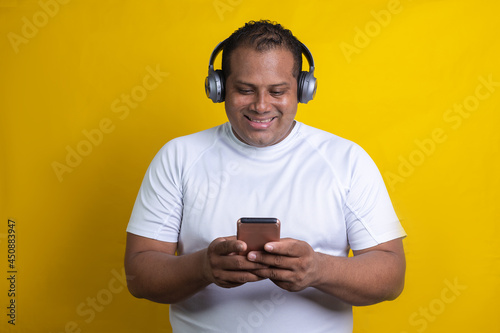 Hispanic man listening to music with headphones and cell phone, on yellow background