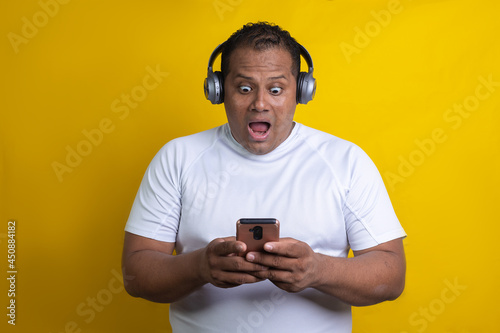 Hispanic man listening to music with headphones and cell phone amazed, in yellow background