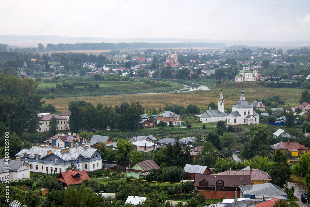 Obraz premium Panoramic top view of the historic old town of Suzdal with the roofs of houses and green foliage of trees on a cloudy summer day