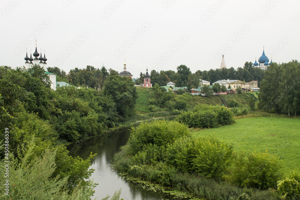 panoramic view of the old town with historical wooden houses and an Orthodox church among the green foliage of trees on the river bank with a reflection on a cloudy day in Suzdal Russia