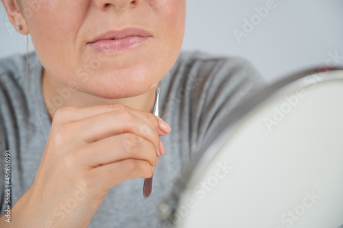 Caucasian woman looks in the mirror and removes hair on her chin herself with the help of tweezers. Hirsutism.