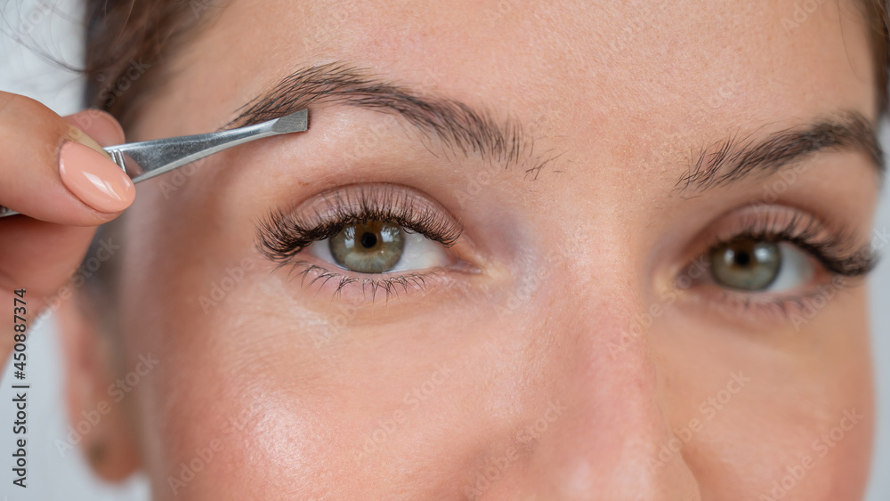 Close-up portrait of a caucasian woman doing eyebrow correction herself with tweezers