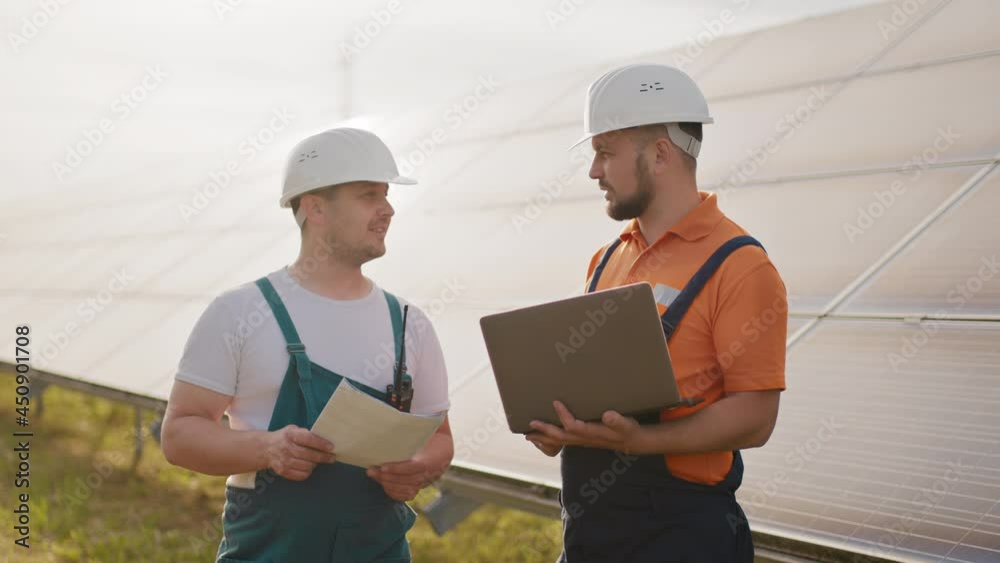 Technician with laptop checking the panels at solar energy installation. Business cooperation. Solar park. Alternative energy concept. Clean energy. Two workers in a uniform and hard hat Technician with laptop checking the panels at solar energy installation. Business cooperation. Solar park. Alternative energy concept. Clean energy. Two workers in a uniform and hard hat