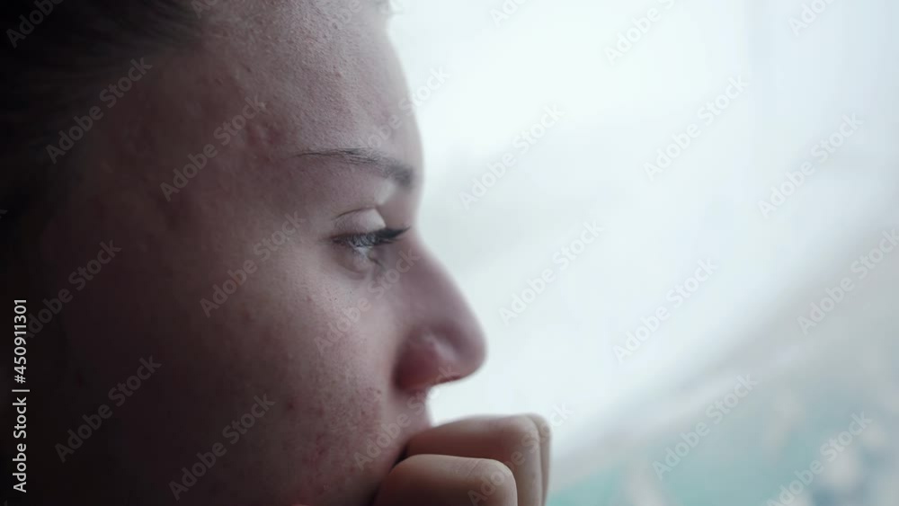 Sad young woman looking out the window at home, sitting alone and watching the snow. Self isolation and winter depression concept