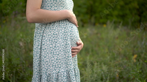 A young pregnant woman stands and holds her belly with her hands. Girl in a dress in nature.