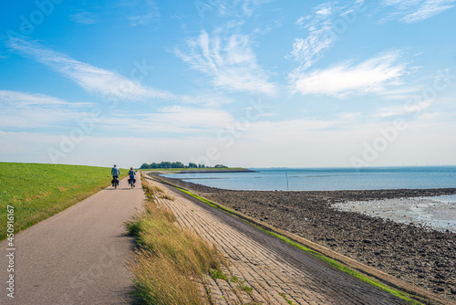 A man and a woman are cycling on a path at the bottom of a Dutch dike and along the Oosterschelde. The dike is reinforced with stones and partly covered with green algae. It is summertime now. photo
