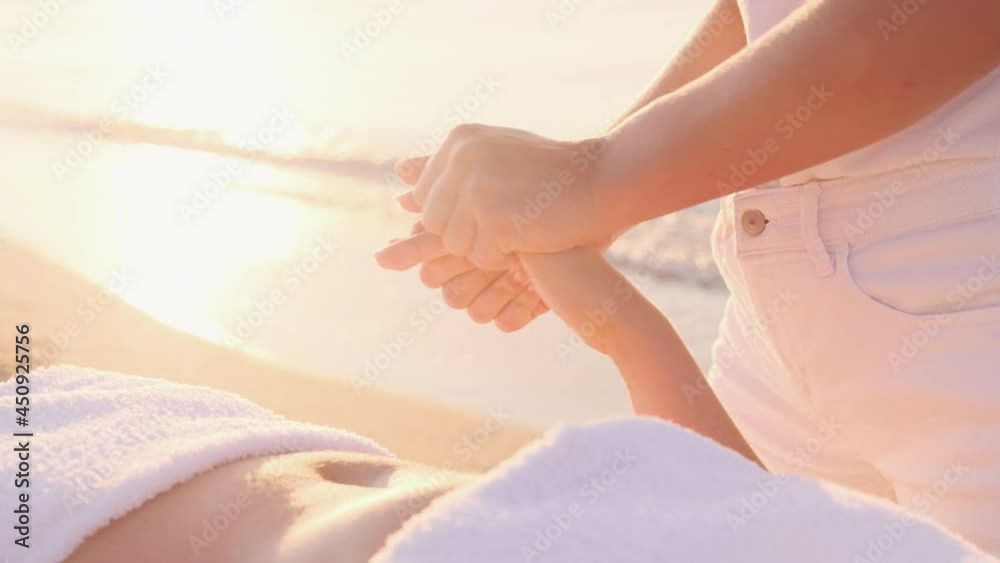 A young masseuse makes a hand massage to a woman on the seashore at sunrise. The rays of the sun through the hands. Massage at the resort. The woman is covered with a white towel.