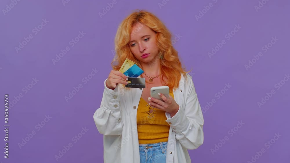 Sincere redhead lovely girl 20s years old in white shirt using credit bank cards and smartphone while transferring money purchases online shopping. Young adult woman isolated on purple wall background Sincere redhead lovely girl 20s years old in white shirt using credit bank cards and smartphone while transferring money purchases online shopping. Young adult woman isolated on purple wall background