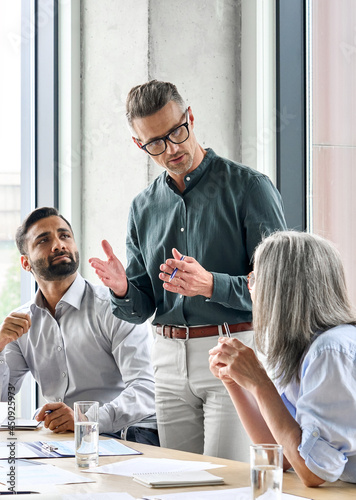Mature businessman leader mentor talking to diverse colleagues team listening to caucasian ceo. Multicultural professionals project managers group negotiating in boardroom at meeting. Vertical.