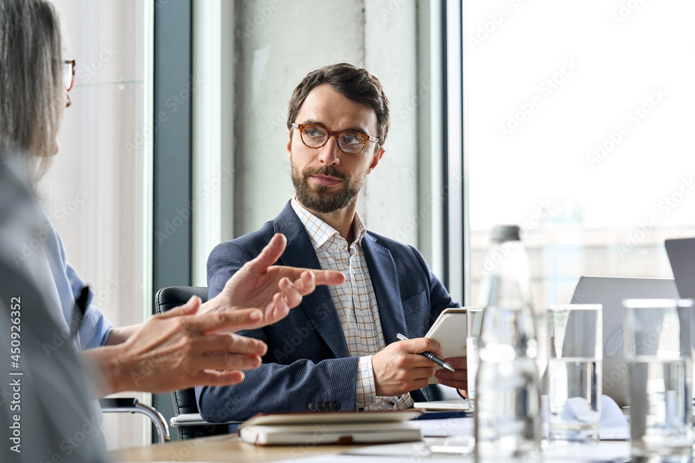 Serious Caucasian bearded male ceo wearing glasses looking at female manager controlling discussing corporation project report at table. Executive managers leaders working together in modern office. Serious Caucasian bearded male ceo wearing glasses looking at female manager controlling discussing corporation project report at table. Executive managers leaders working together in modern office.