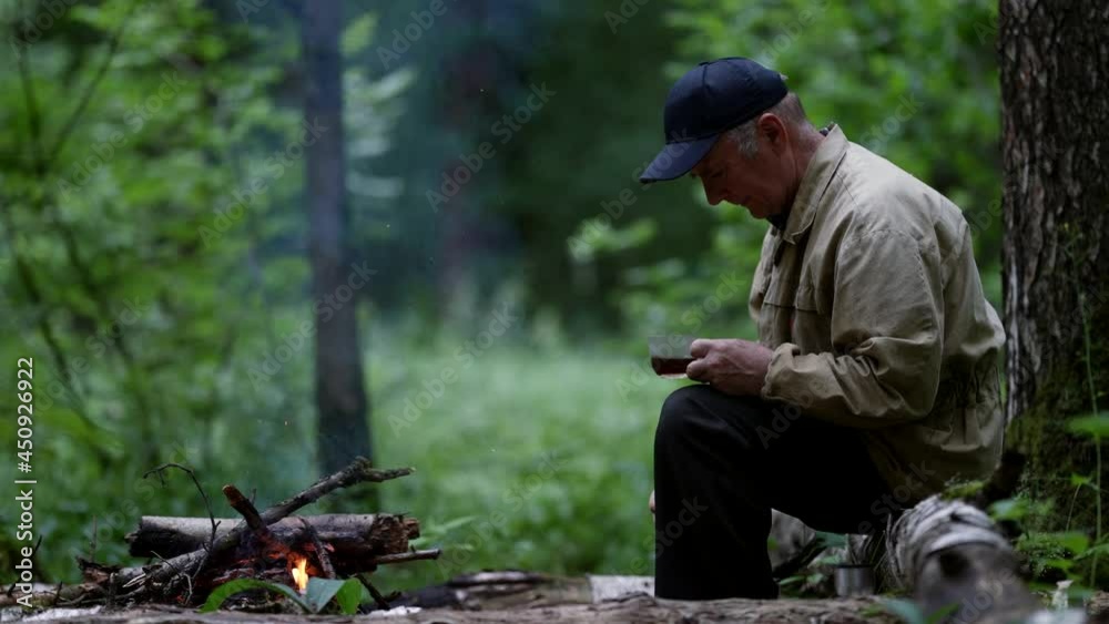 old man is trekking alone in forest, resting near campfire and drinking hot tea old man is trekking alone in forest, resting near campfire and drinking hot tea