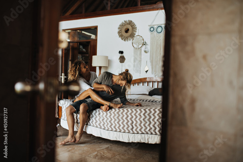 On photo taken through open doors, loving couple is having fun sitting on big bed