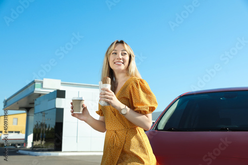 Beautiful young woman with coffee and hot dog near car at gas station