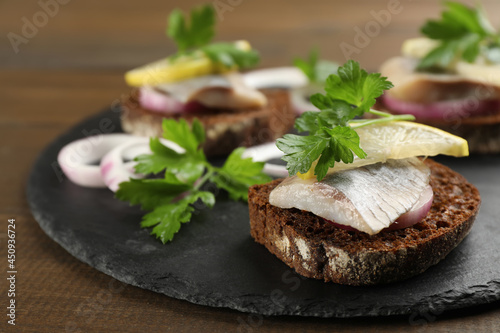 Delicious sandwiches with salted herring, onion rings, parsley and lemon on wooden table, closeup